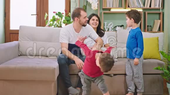 他们微笑着与两个孩子和有魅力的父母在沙发上享受家中时光的大而快乐的年轻家庭视频的预览图