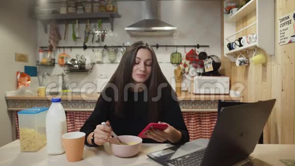 年轻女性在家里的厨房里使用笔记本电脑智能手机和玉米片视频的预览图