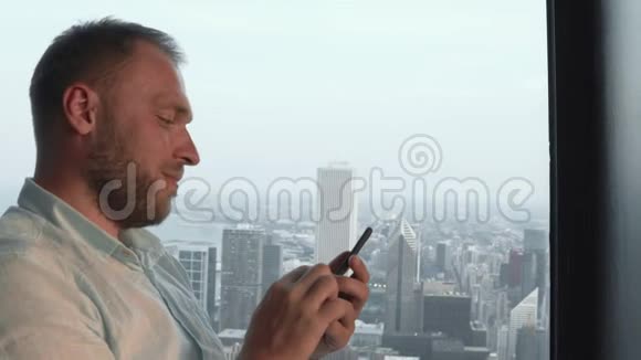 年轻的帅哥站在摩天大楼的窗户附近用智能手机美国芝加哥全景视频的预览图