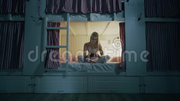 年轻漂亮的女人坐在宿舍的床上晚上用智能手机视频的预览图
