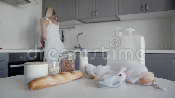 厨房木制桌子上有面包燕麦片荞麦牛奶蔬菜和水果的未包装购物袋妇女视频的预览图