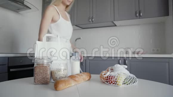 金发女人把面包燕麦片牛奶蔬菜从桌子移到架子上零废家和生态袋生态购物视频的预览图