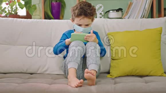 有魅力的小男孩坐在沙发上玩智能手机他戴着保护面具集中精力玩视频的预览图