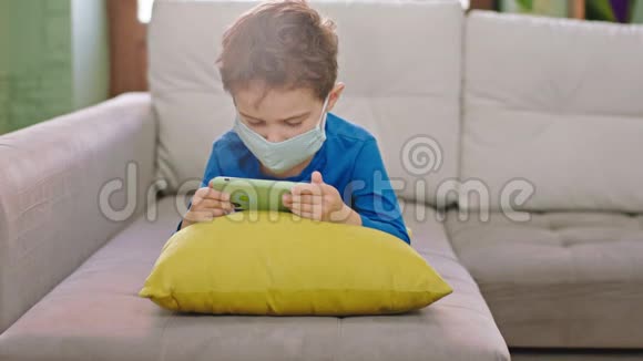 一个小男孩带着智能手机呆在家里戴着防护面具在沙发上玩一个集中的游戏的肖像视频的预览图