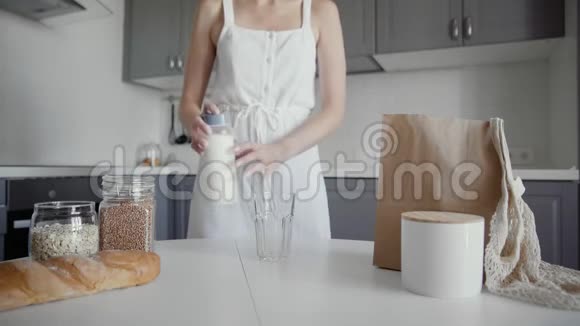穿着白色连衣裙的女人把牛奶从老式的瓶子倒进桌子上的玻璃杯里这是一个面目全非的女人视频的预览图