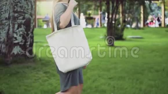 少女侧景手持白色纺织生态袋漫步夏日公园视频的预览图