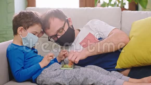 一位父亲带着他的小孩子呆在家里隔离他们戴着防护装置在智能手机上玩视频的预览图