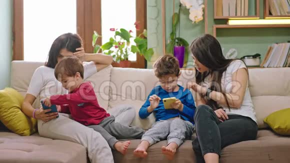 两个年轻的母亲呆在家里和他们的孩子聊天而坐在沙发上的孩子使用智能手机视频的预览图