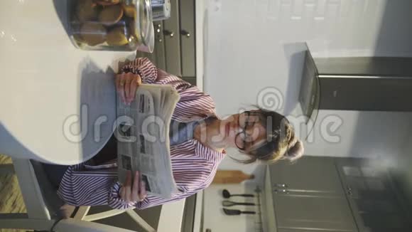垂直视频年轻美丽的黑发女人吃饼干和看报纸在家里厨房视频的预览图