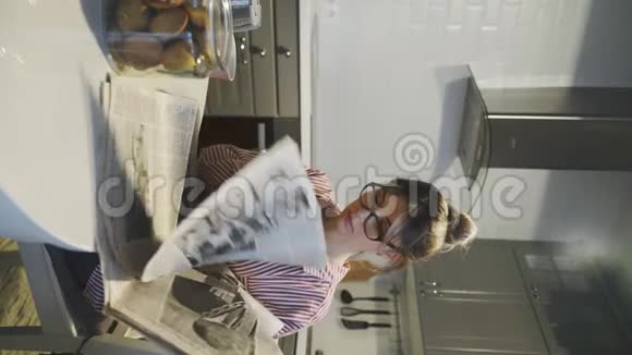 带眼镜的年轻漂亮黑发女人在家厨房里看报纸的垂直视频视频的预览图