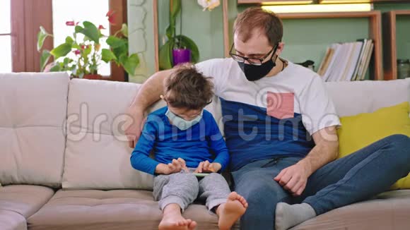 检疫时间在家年轻的父亲和他的小儿子带着保护面具坐在沙发上玩视频的预览图