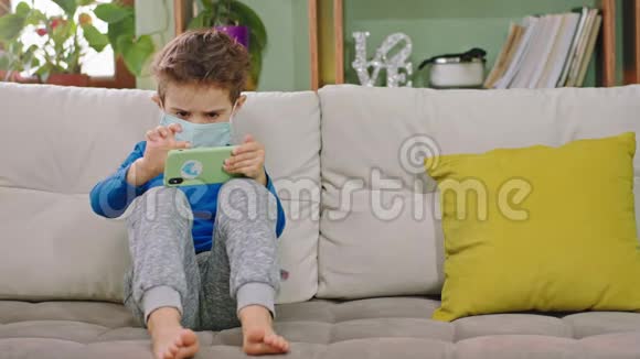 在家里的隔离集中了一个戴着防护面罩的小男孩坐在智能手机上玩游戏视频的预览图