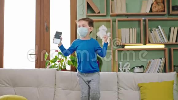 在沙发上快乐的跳跃男孩戴着一个防护面具他呆在家里隔离手里拿着他的智能手机视频的预览图