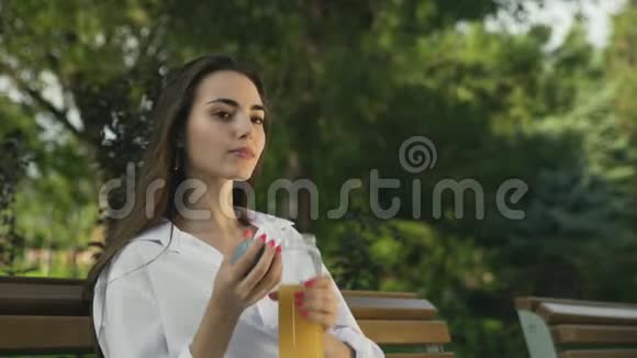 年轻漂亮的女孩子在夏天的公园里从瓶子里喝橙汁视频的预览图