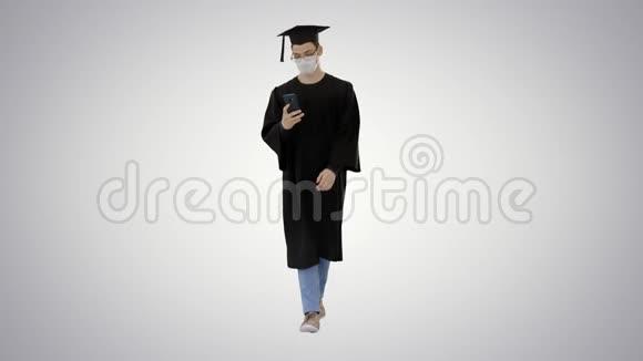 带毕业礼服的年轻人戴着医用面具在渐变背景下使用智能手机行走视频的预览图
