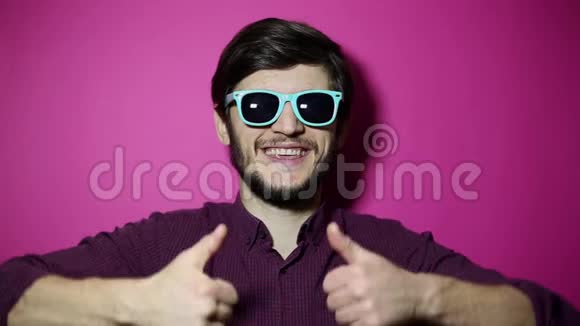 年轻快乐男子的肖像竖起大拇指戴着蓝色太阳镜背景是粉红色视频的预览图