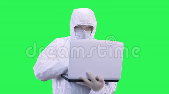 一个戴着绿色背景保护服的戴口罩和眼镜的男人专心地把数据输入笔记本电脑视频的预览图