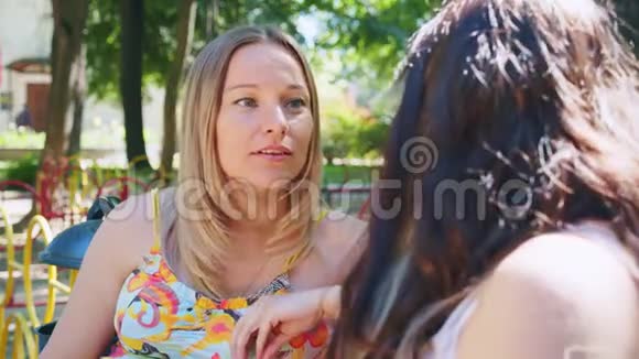 两个美丽的年轻女子金发和黑发在夏季公园的情感交谈视频的预览图