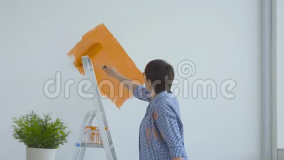 平面改造概念快乐中年妇女用油漆滚筒画白色墙壁橙色油漆视频的预览图