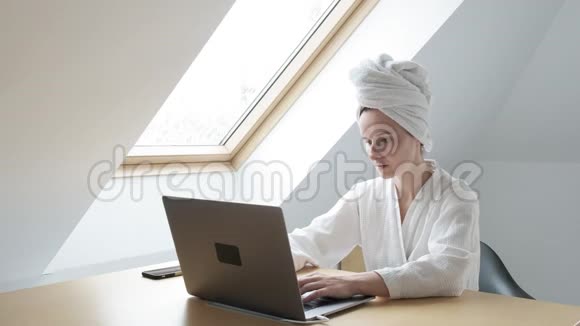 身穿白色浴袍头戴毛巾的年轻女子在家中手提电脑远程工作视频的预览图