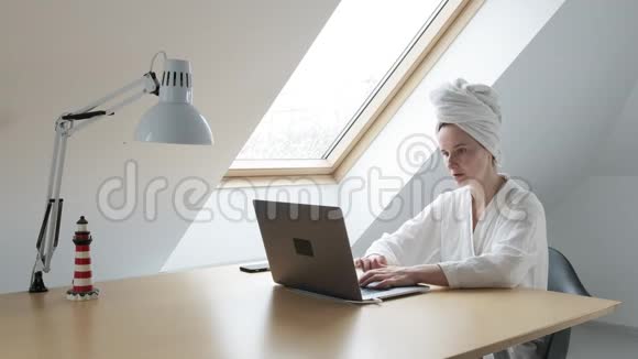 身穿白色浴袍头戴毛巾的年轻女子在家中手提电脑远程工作视频的预览图