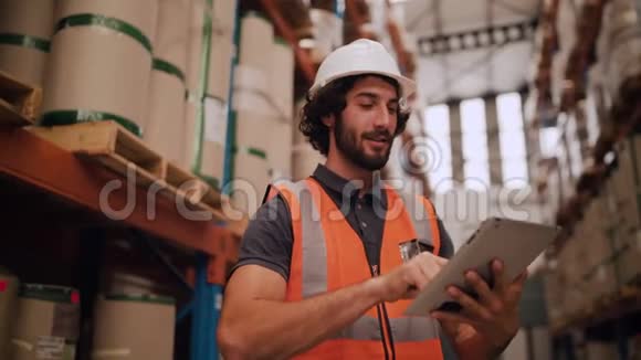 戴着白色安全帽的年轻人在大仓库里用数码平板电脑背景是一堆货物视频的预览图