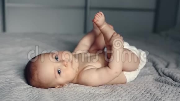 可爱的笑着的白种人6个月大的小男孩抱着他的脚躺在床上看着相机视频的预览图