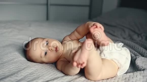 可爱的笑着的白种人6个月大的小男孩抱着他的脚躺在床上看着相机视频的预览图