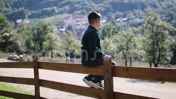 一个8岁的小男孩坐在木栅栏上看着群山视频的预览图