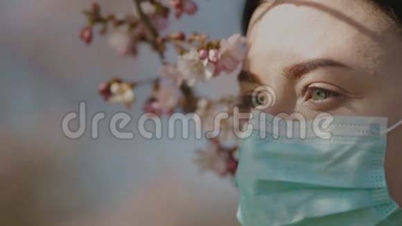 美丽悲伤的年轻女子绿色的眼睛戴着外科口罩看着远处慢镜头拍摄樱花花视频的预览图