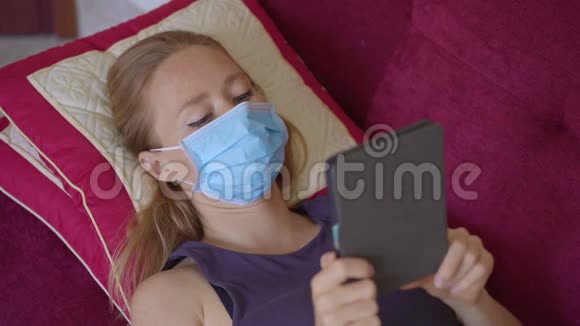 一位戴着医用口罩的年轻女子躺在沙发上看着一本电子书她在家吃冠状病毒视频的预览图