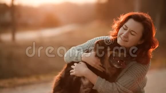 可爱的红发女人在模糊温暖的背景下在日落时表现出舌头和拥抱并与卷曲狗玩耍慢动作视频的预览图