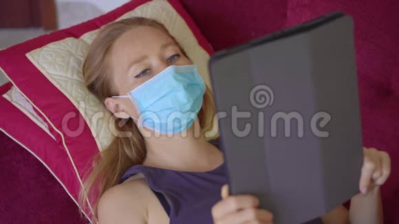 一位戴着医用口罩的年轻女子躺在沙发上看着一本电子书她在家吃冠状病毒视频的预览图