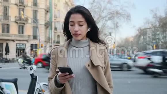 漂亮的年轻女人在街上散步时用手机和无线耳机听音乐视频的预览图