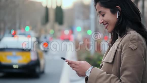 漂亮的年轻女人在街上用手机发短信用无线耳机听音乐视频的预览图