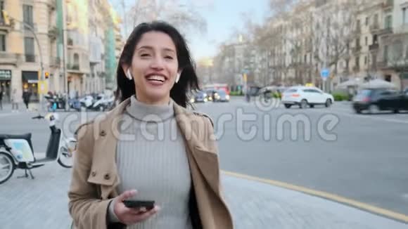 漂亮的年轻女人在街上散步时用无线耳机在手机上说话视频的预览图