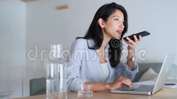 优雅的年轻商务女性在办公室用笔记本电脑工作时用手机说话视频的预览图