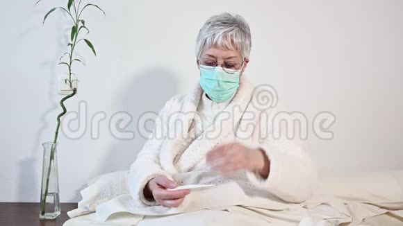 冠状病毒生病的老年妇女戴着防护面罩拿着温度计坐在床上让老成女人心烦意乱感冒了视频的预览图