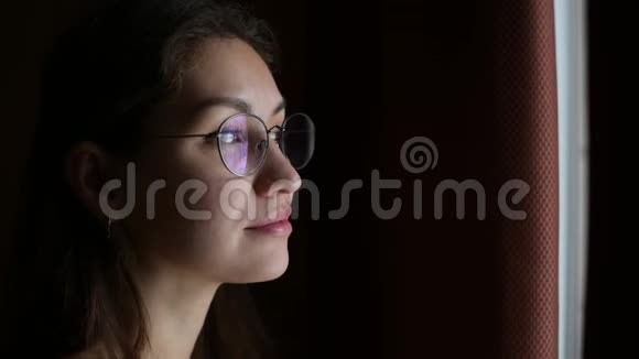 戴眼镜的年轻女子从一间昏暗的房间里向窗外沉思地看着视频的预览图