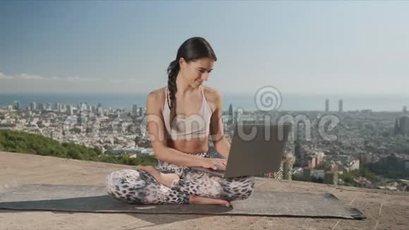 巴塞罗那市的女孩用笔记本电脑瑜伽女人坐在垫子上的莲花姿势视频的预览图