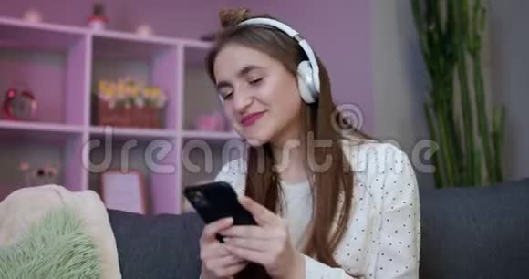 耳机里的快乐女孩是在沙发上跳舞用智能手机触摸屏幕现代技术概念视频的预览图