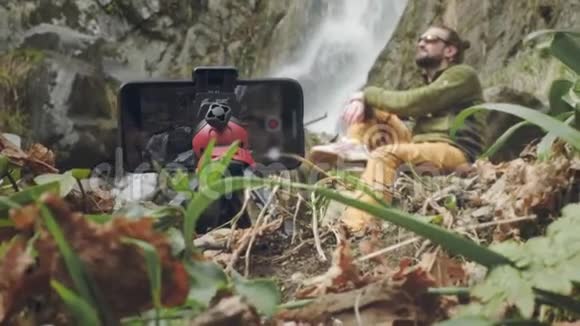年轻的男性旅行者博主坐着享受美丽的瀑布在山里远足一个徒步旅行者坐在视频的预览图