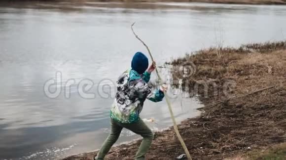 一个男孩从河岸把一个长树枝扔进水里慢镜头图像视频的预览图