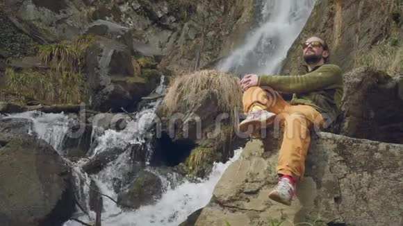 年轻的男性旅行者坐着享受美丽的瀑布在山里远足一个徒步旅行者坐在瀑布旁视频的预览图