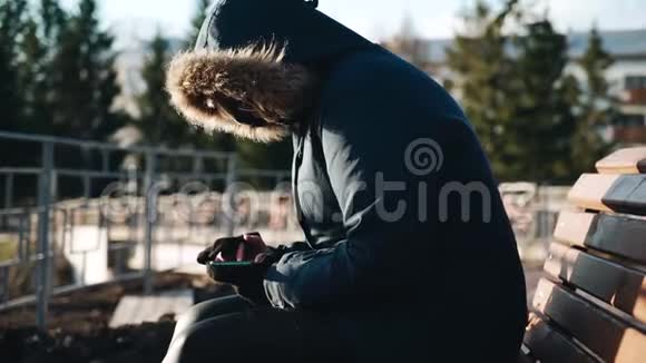 一个穿着夹克坐在长凳上手里拿着智能手机的女人周围没有人视频的预览图