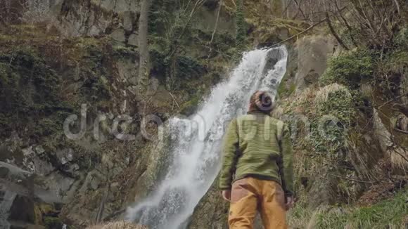 年轻的男性旅行者喜欢美丽的瀑布在山里远足徒步旅行者跑到瀑布前举起视频的预览图