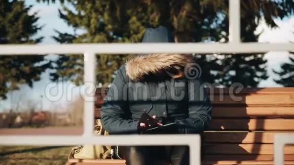 日落时分一个穿着夹克衫头罩的女人坐在公园的长凳上视频的预览图