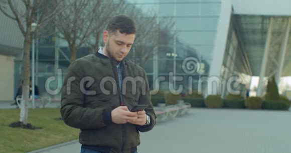 带着智能手机走在现代办公中心附近的男子肖像在社交网络上和朋友聊天看视频的预览图