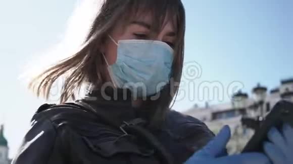 在冠状病毒流行期间年轻女性戴着外科口罩和手套在户外使用智能手机视频的预览图