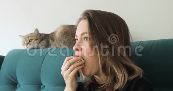 年轻女子正坐在沙发上吃爆米花和猫一起看电视视频的预览图